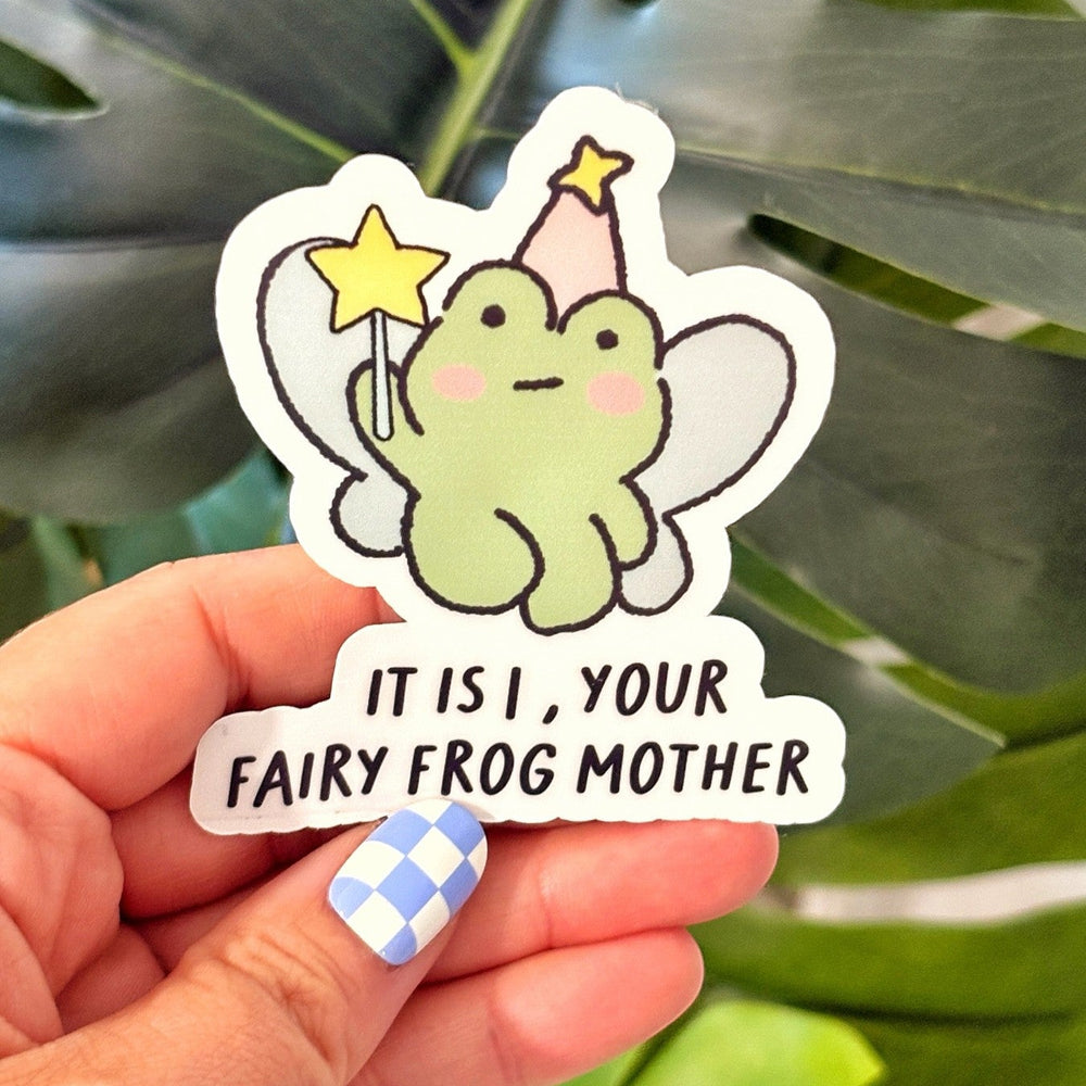 Hand holding a glossy vinyl sticker of a cute green frog with wings, a party hat, and a magic wand. The sticker text reads “It is I, your fairy frog mother.” Purple storage bins are blurred in the background.