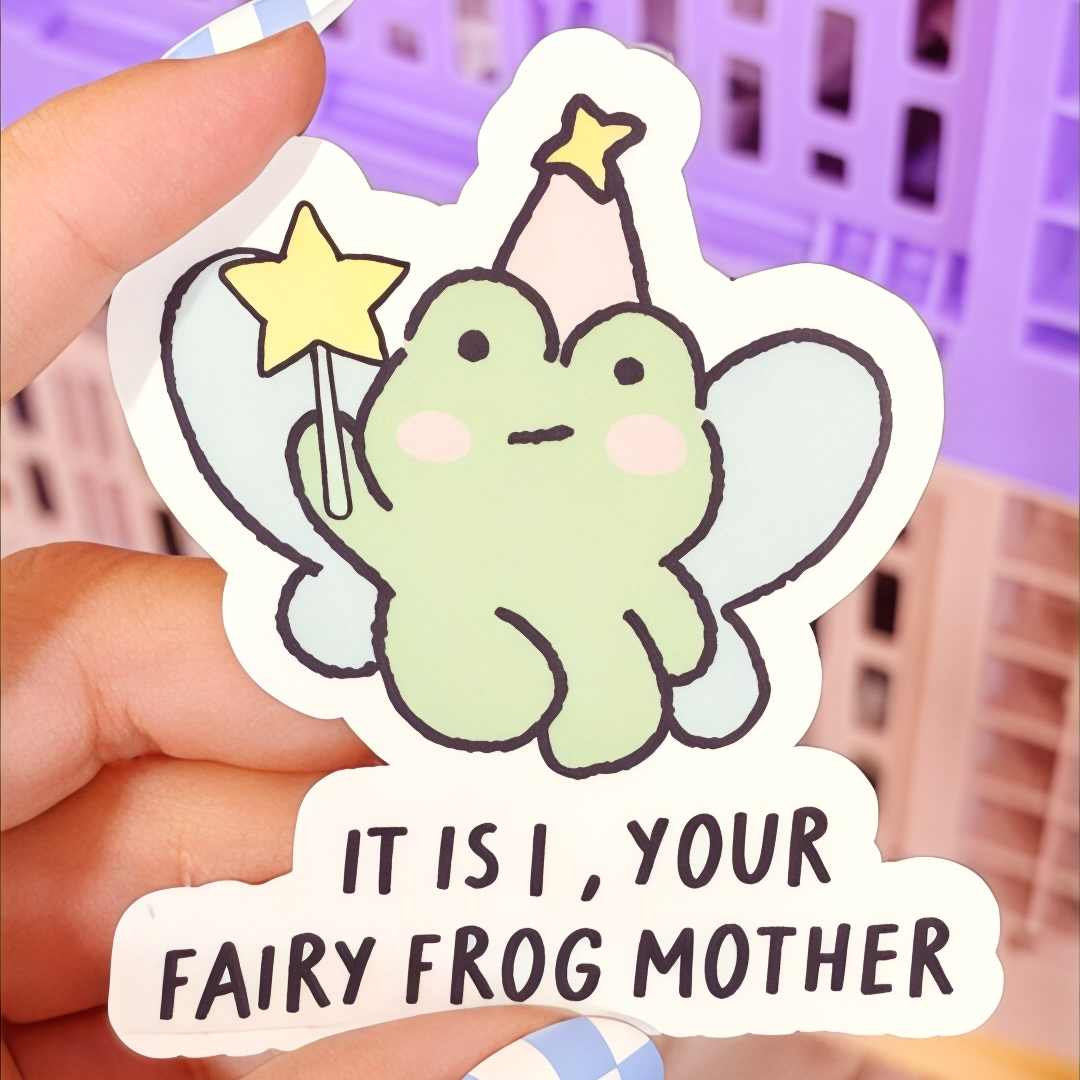 Hand holding a glossy vinyl sticker of a cute green frog with wings, a party hat, and a magic wand. The sticker text reads “It is I, your fairy frog mother.” Purple storage bins are blurred in the background.
