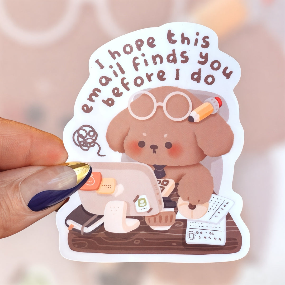 A cute matte vinyl sticker featuring a brown puppy wearing round glasses and a pencil behind its ear, sitting at a desk with a computer, coffee cup, and papers. The text above reads “I hope this email finds you before I do.” The design has a cozy, pastel office aesthetic.